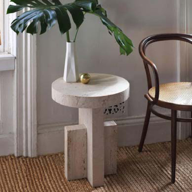 Pale stone side table with a thick round top and intersecting slab legs beside a chair