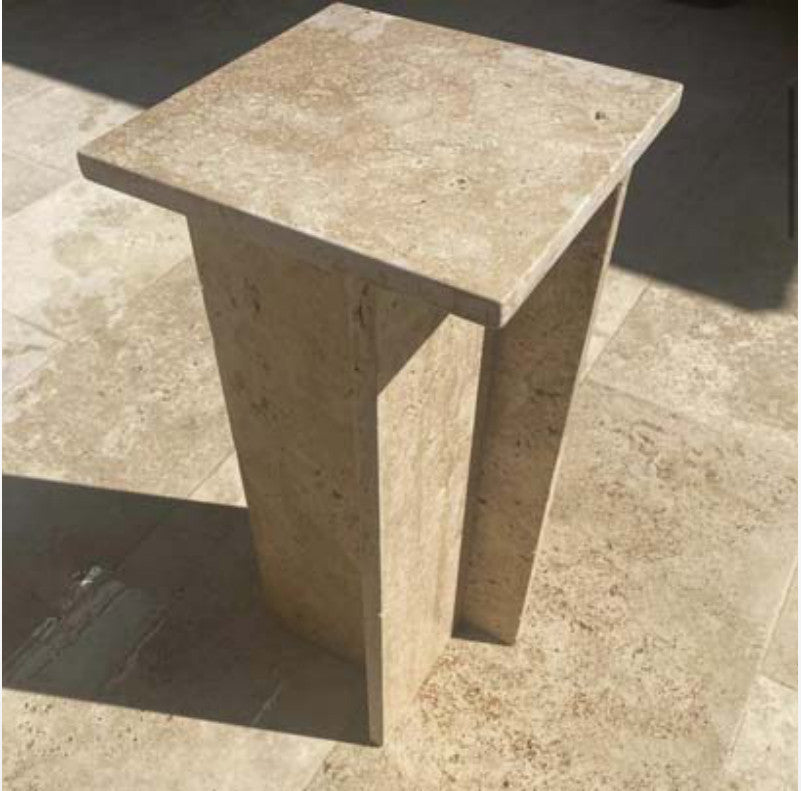 Beige stone side table with a square top and two splayed slab supports in sunlight