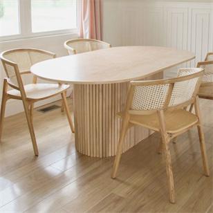 Oval pale stone dining table with a fluted pedestal and wooden chairs beside a window
