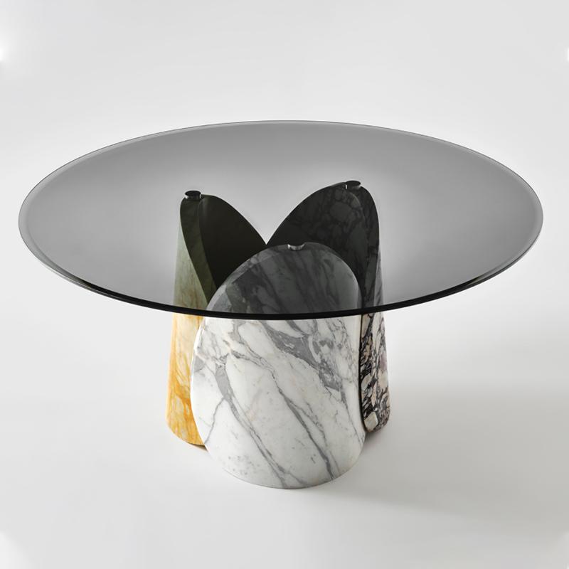 Round dining table with a transparent top and intersecting stone supports, three-quarter view