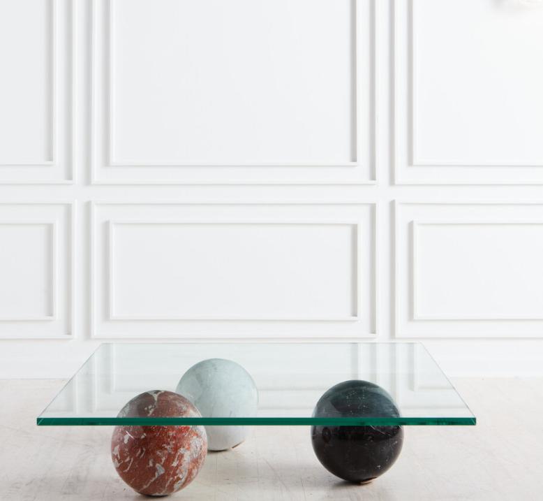 Rectangular transparent tabletop resting on coloured stone spheres in front of a panelled wall
